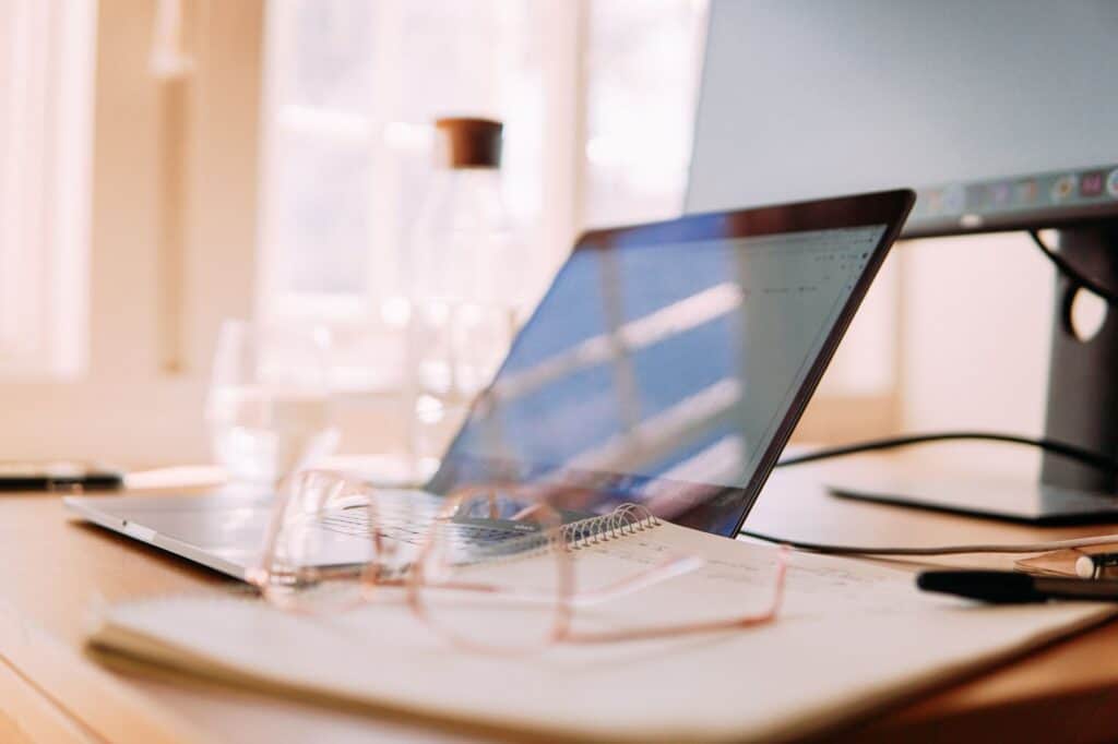 A well-lit home office setup featuring a laptop, monitor, and glasses, ideal for remote work.